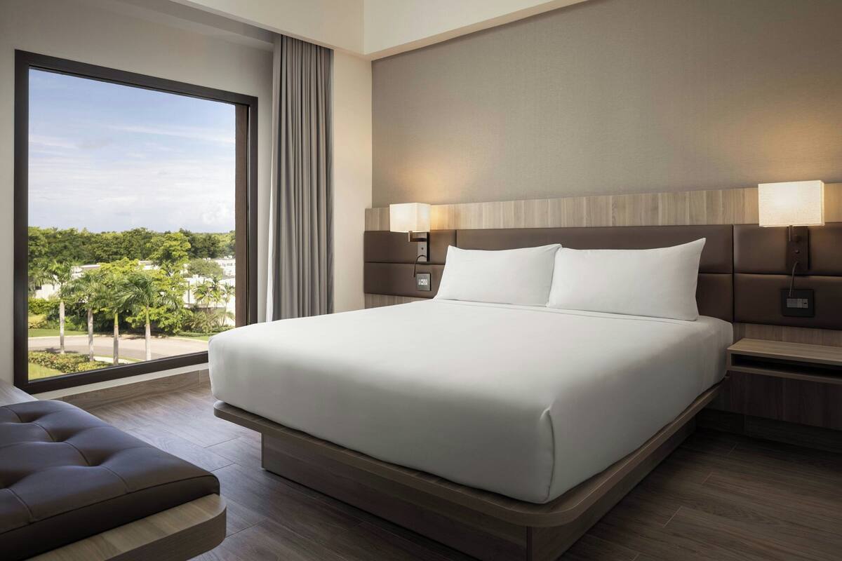 A modern hotel room with a large bed, bedside tables, and a view of greenery outside.