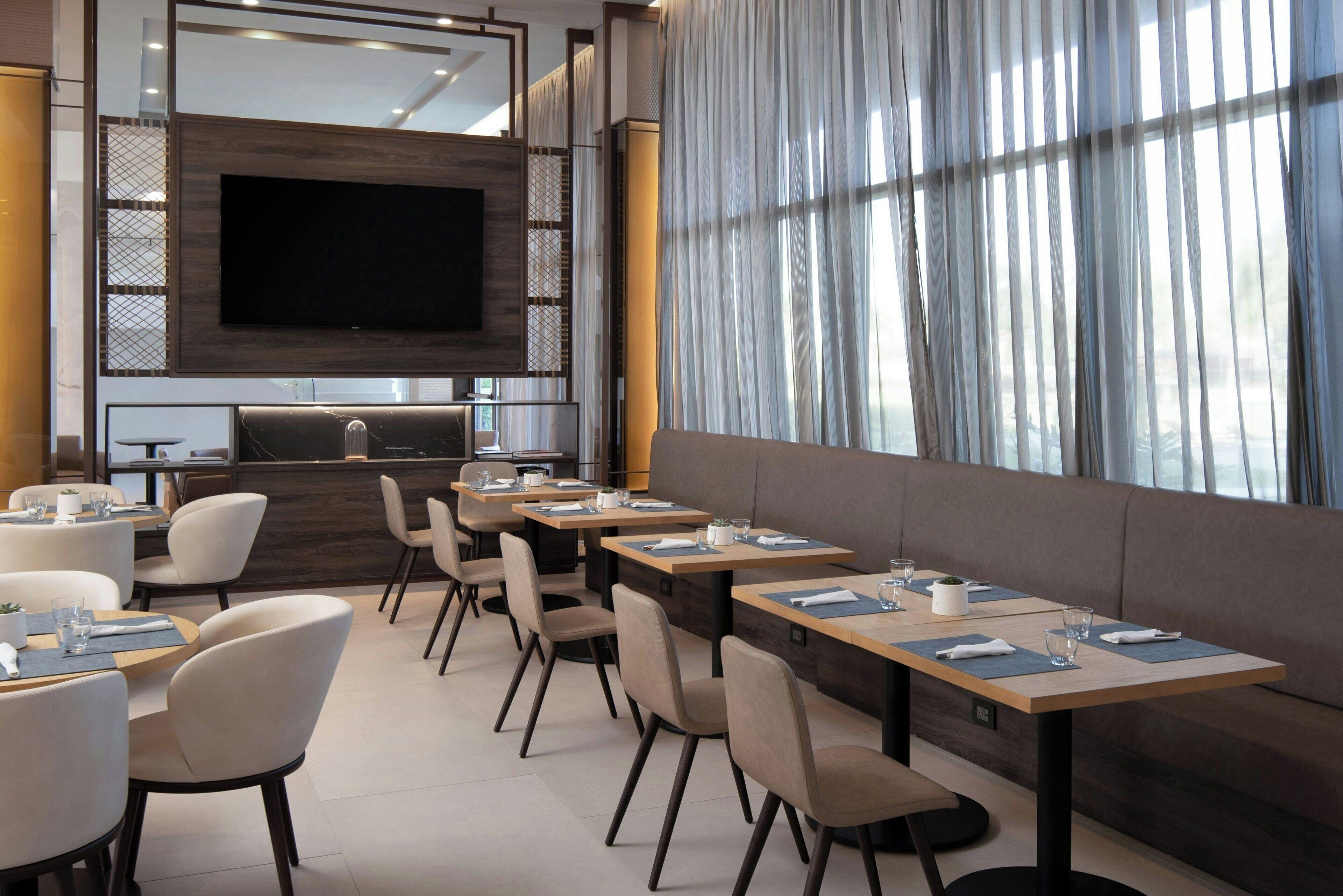 A modern hotel dining area with tables and chairs, a large flat-screen TV mounted on the wall, and a large window with sheer curtains.