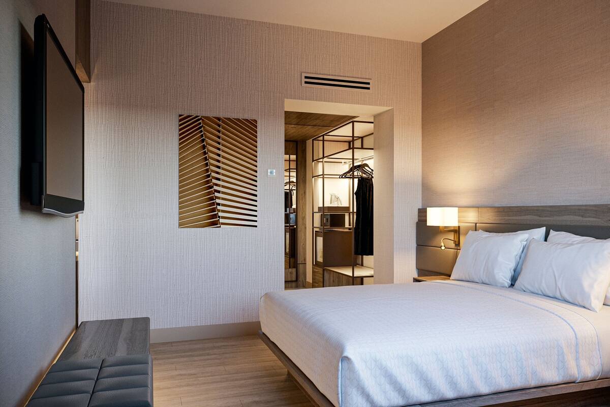A modern hotel room with a large bed, a bench, a TV, and a walk-in closet.