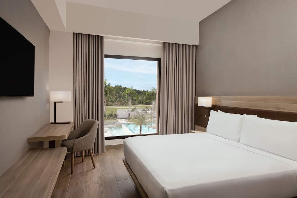 A hotel room with a bed, a desk, a chair, a TV, and a view of a swimming pool and greenery.