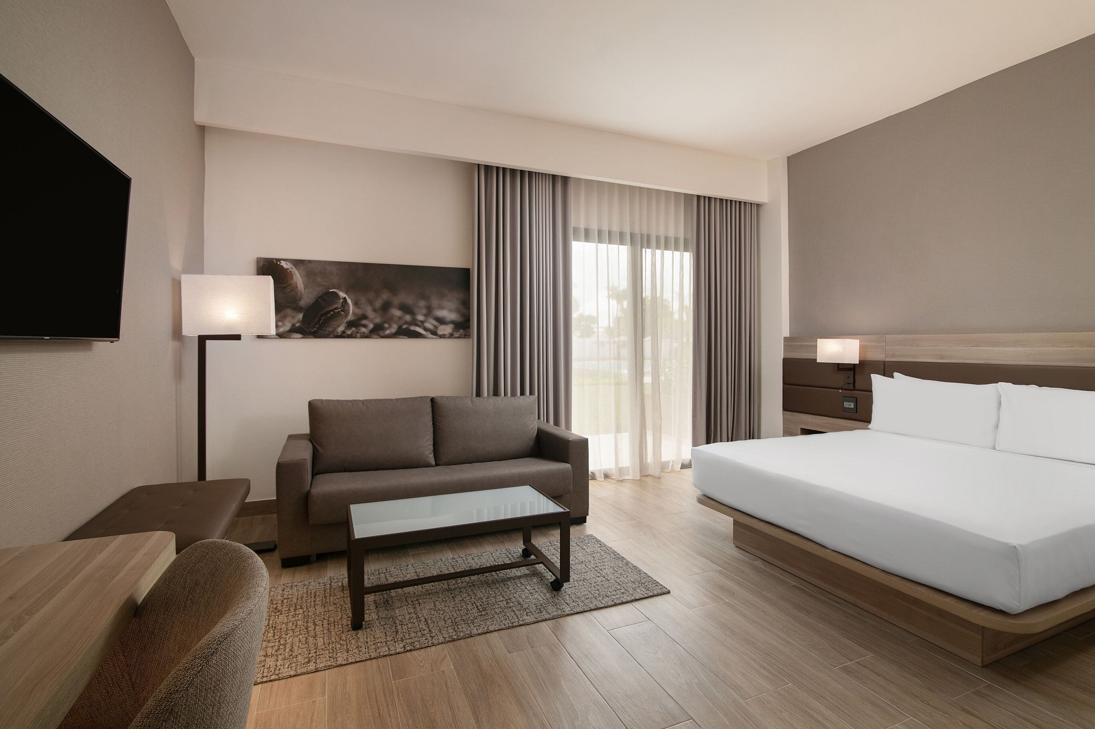 A modern hotel room with a large bed, a sofa, a coffee table, and a flat-screen TV.