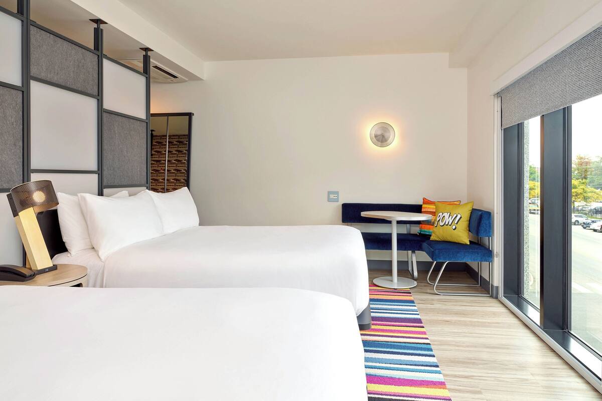 Ett hotellrum med två sängar, ett litet bord, en soffa med en kudde med texten ”POW!”, en färgglad randig matta och ett stort fönster med utsikt över gatan.