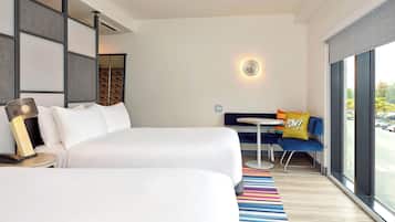Habitación de hotel con dos camas, una mesita, un sofá con un cojín que dice '¡POW!', una alfombra a rayas colorida y un ventanal con vistas a la calle.
