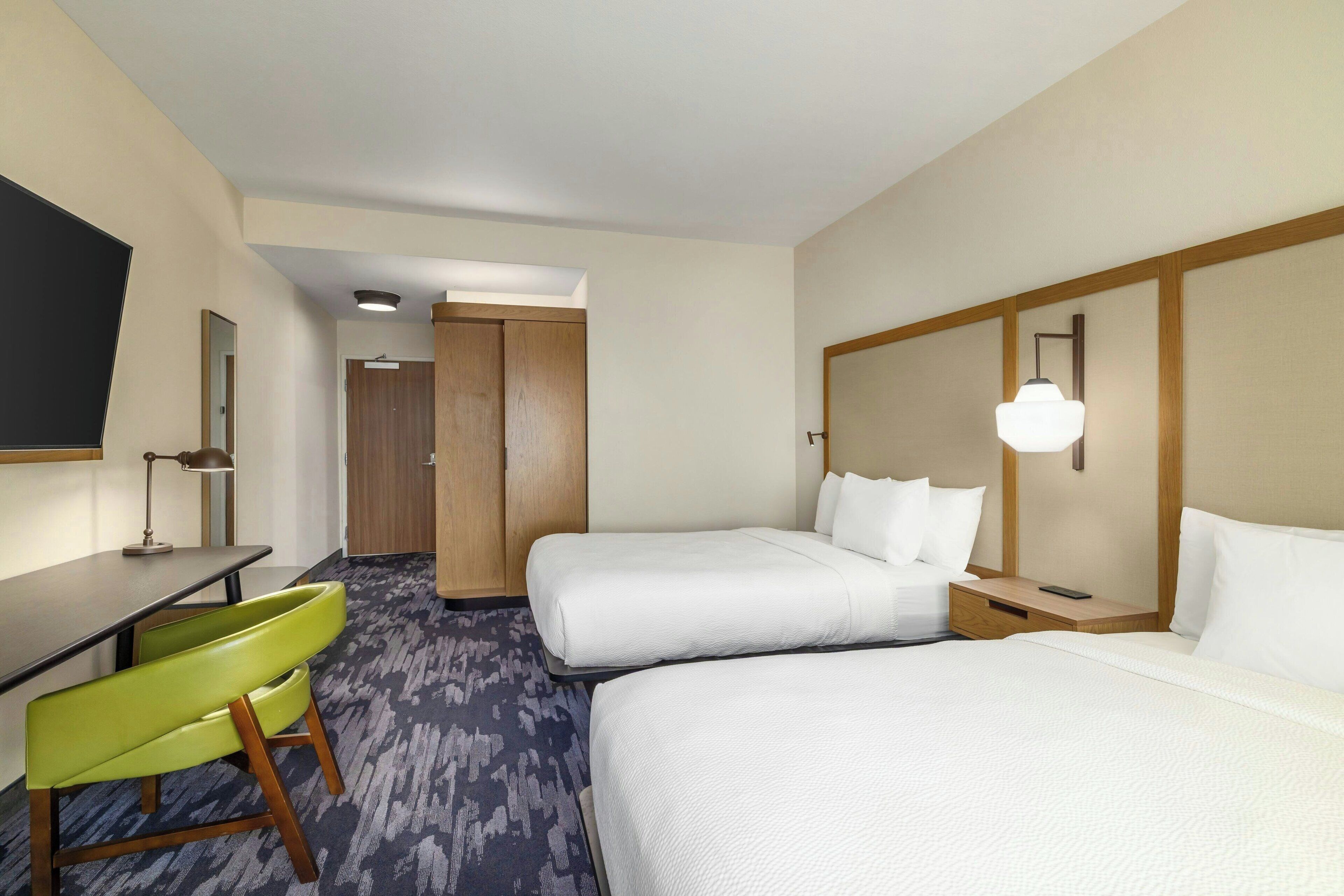 A hotel room with two beds, a desk, a chair, and a TV.