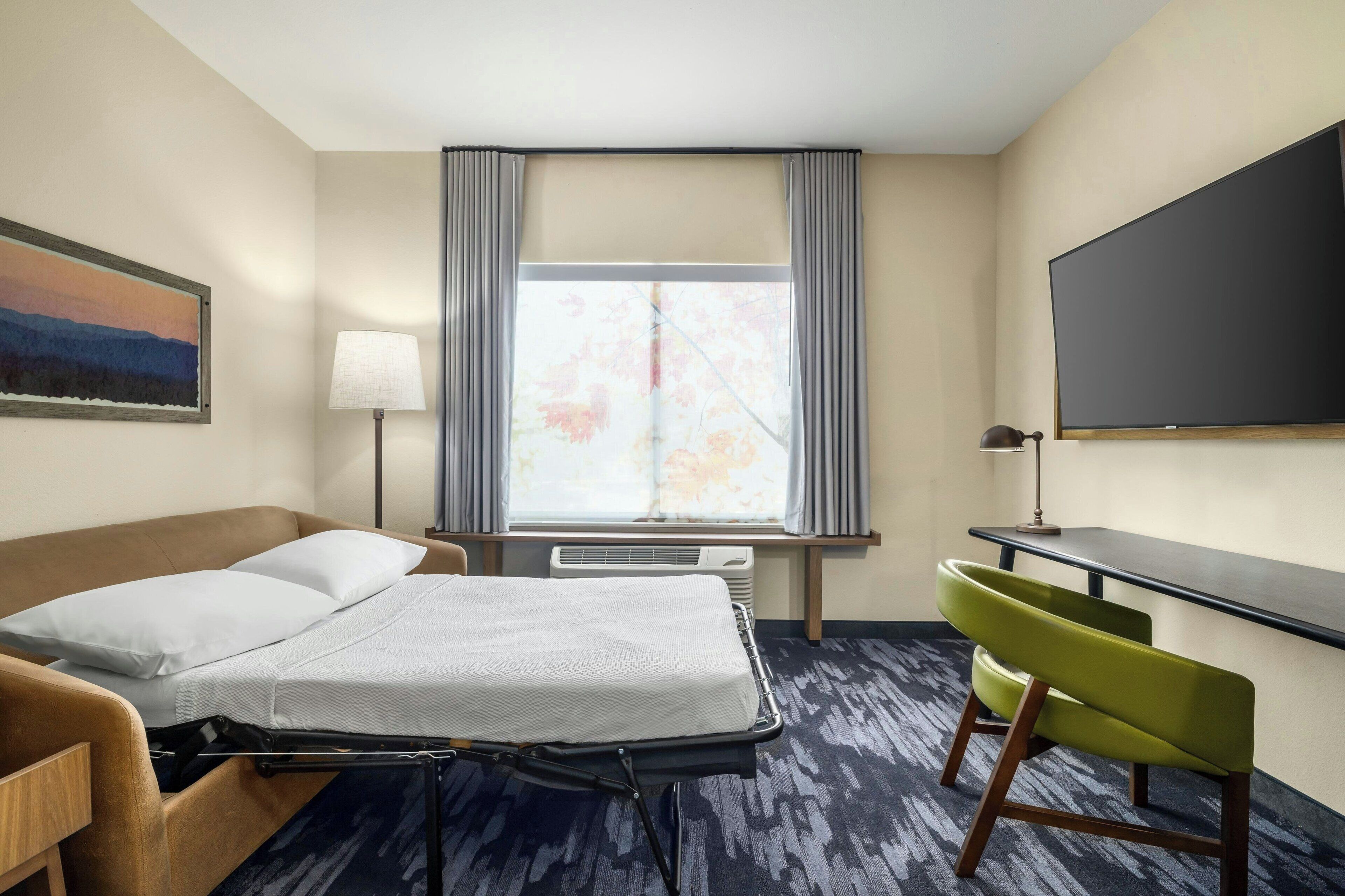 Habitación de hotel con cama, escritorio, silla, ventana grande con cortinas, televisor de pantalla plana y un cuadro en la pared.