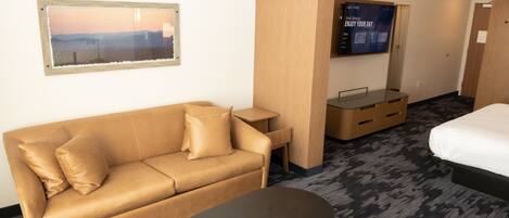 A hotel room with a tan sofa, a small table, a green chair, a bed, and a TV mounted on the wall.