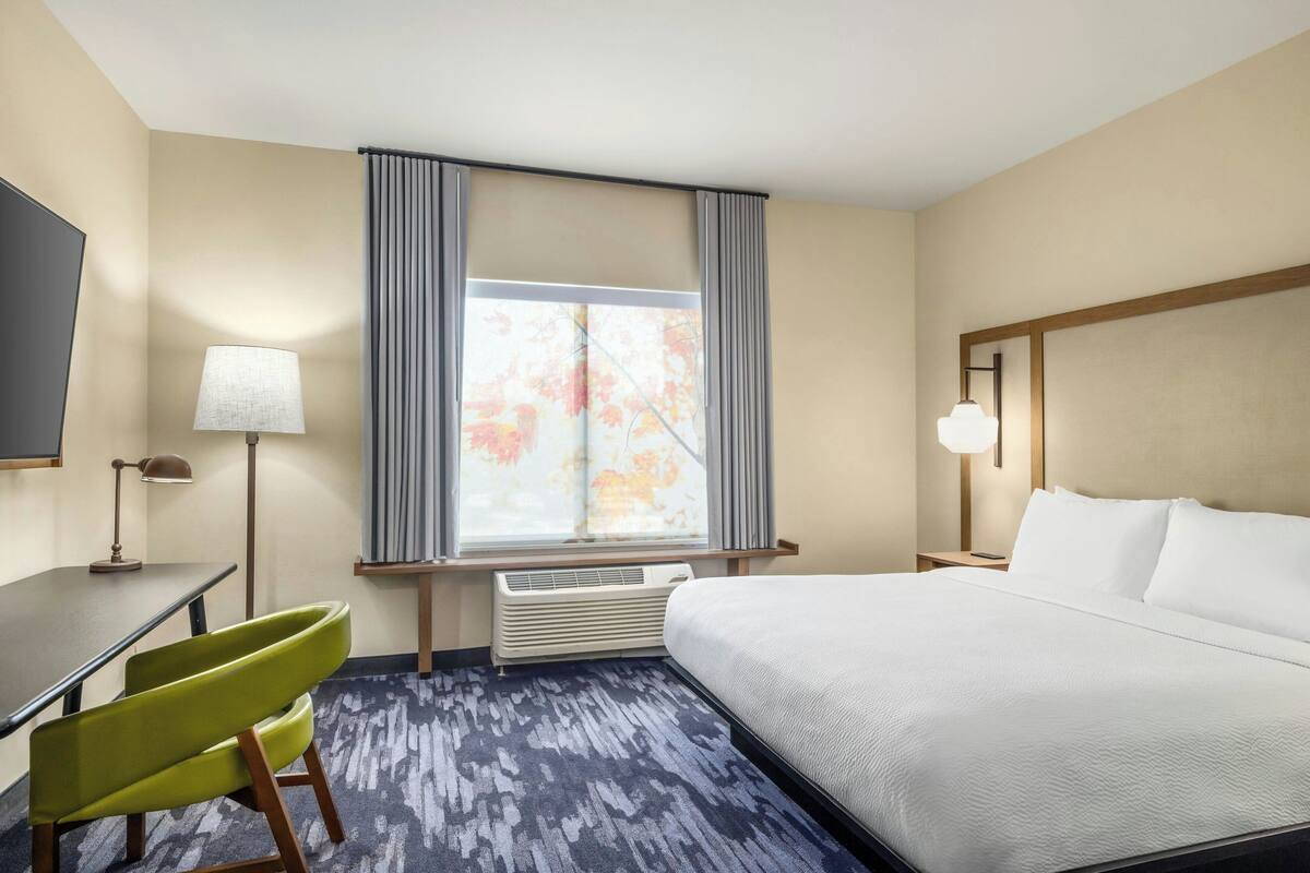 Une chambre d’hôtel équipée d’un lit, d’un bureau, d’une chaise, d’une lampe, d’une fenêtre avec des rideaux et d’un appareil de climatisation.