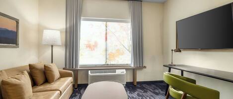 A hotel room with a beige sofa, a small table, a green chair, a large window with curtains, a flat-screen TV, and a wall-mounted air conditioner.