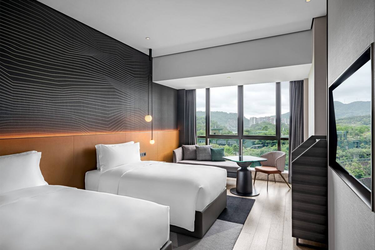 A modern hotel room with two beds, a sofa, a small table, and a view of the cityscape.