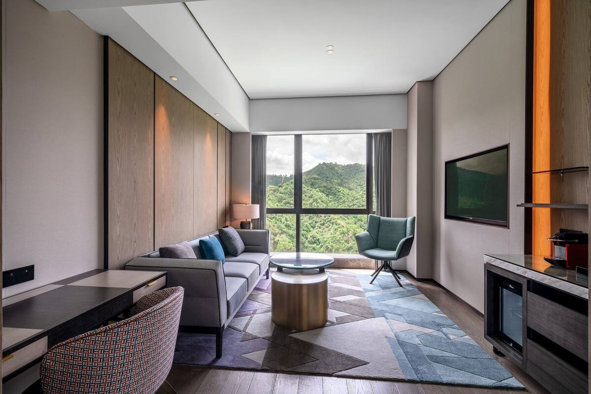 A modern living room with a sofa, a round coffee table, a TV, and a large window with a view of greenery.