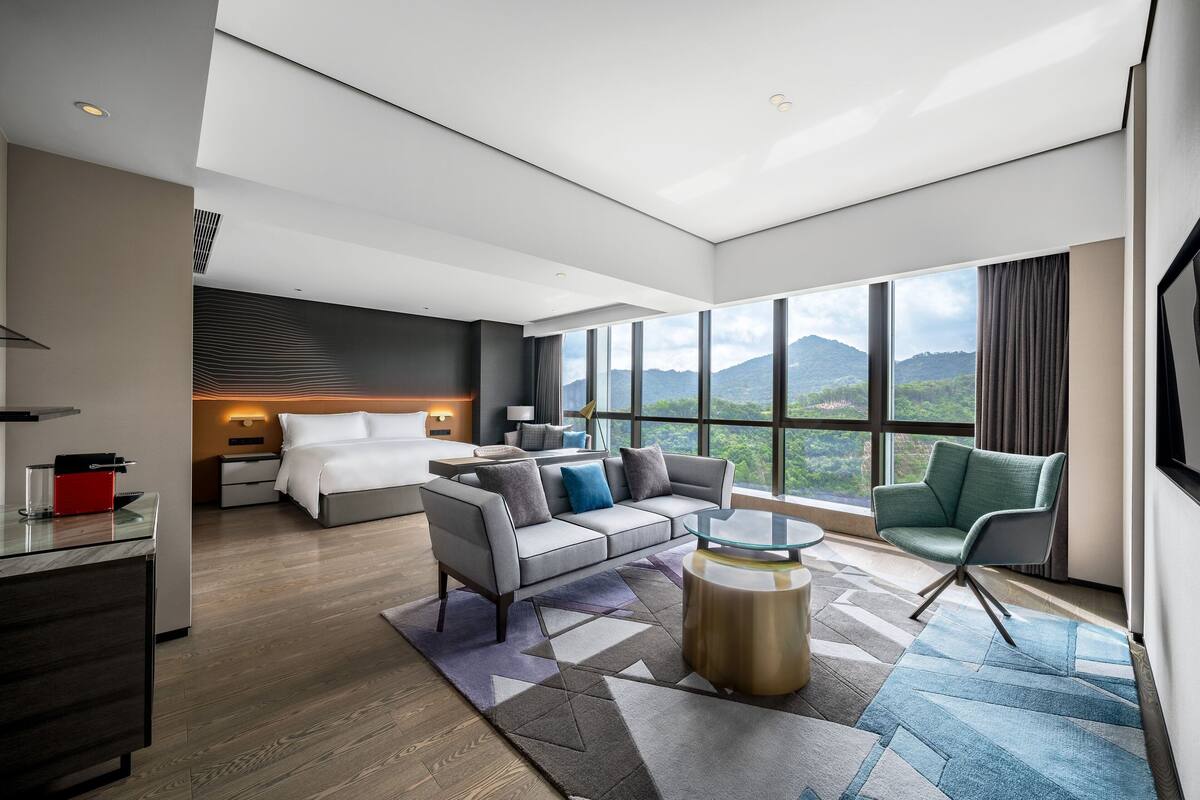A modern hotel room with a large bed, a sofa, a desk, and a chair. There is a large window with a view of mountains.