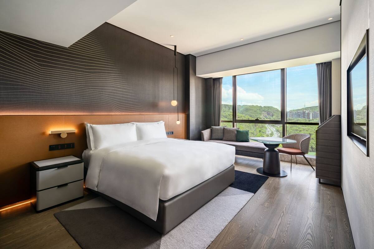 A modern hotel room with a large bed, bedside tables, a seating area with a table, and a view of greenery through large windows.