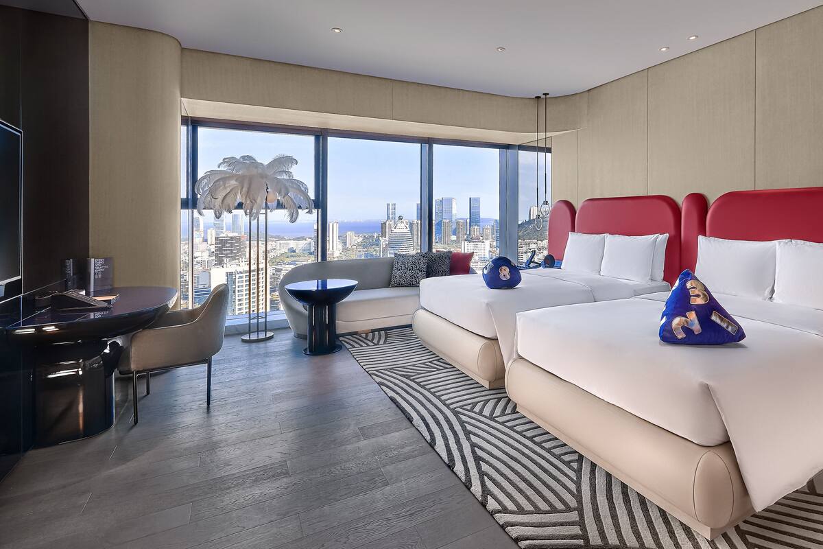 A modern hotel room with a city view, a desk with a chair, a sofa, and two beds with red headboards.