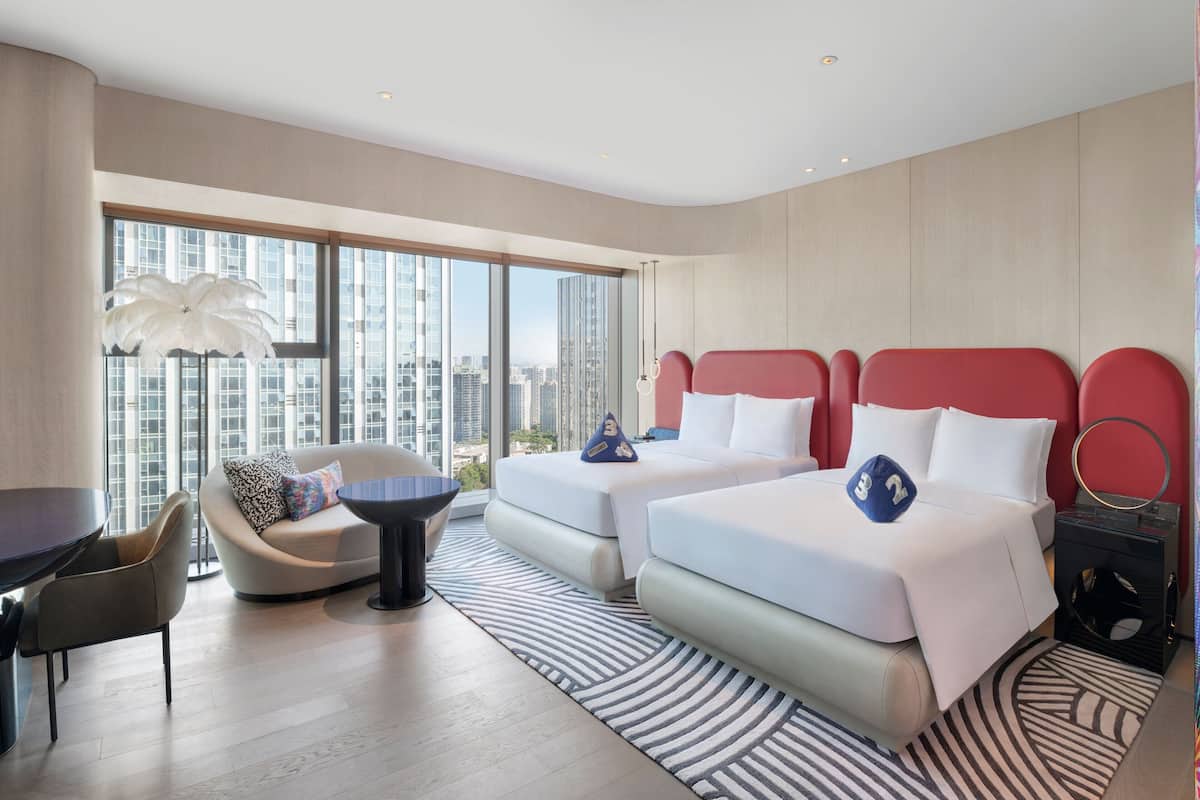 A modern hotel room with two beds, a sofa, a dining table, and a view of the city.
