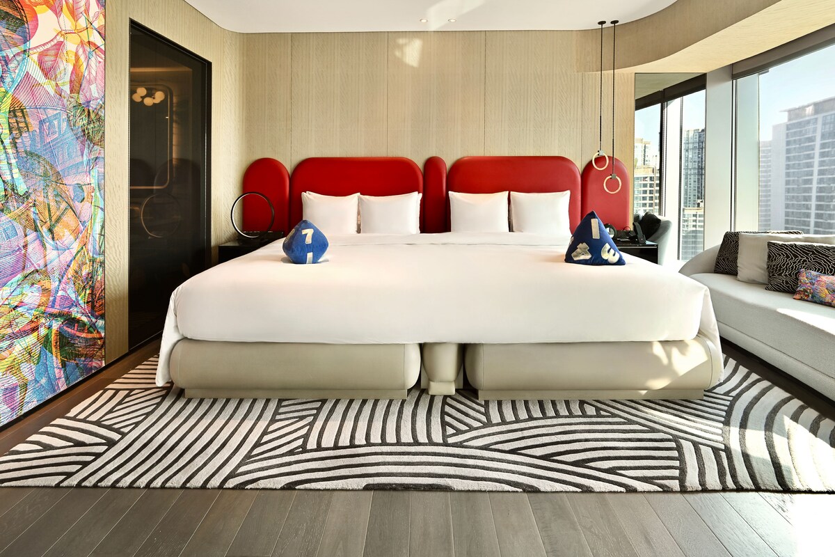 Een moderne hotelkamer met een groot bed, stadszicht en een kleurrijk abstract schilderij.