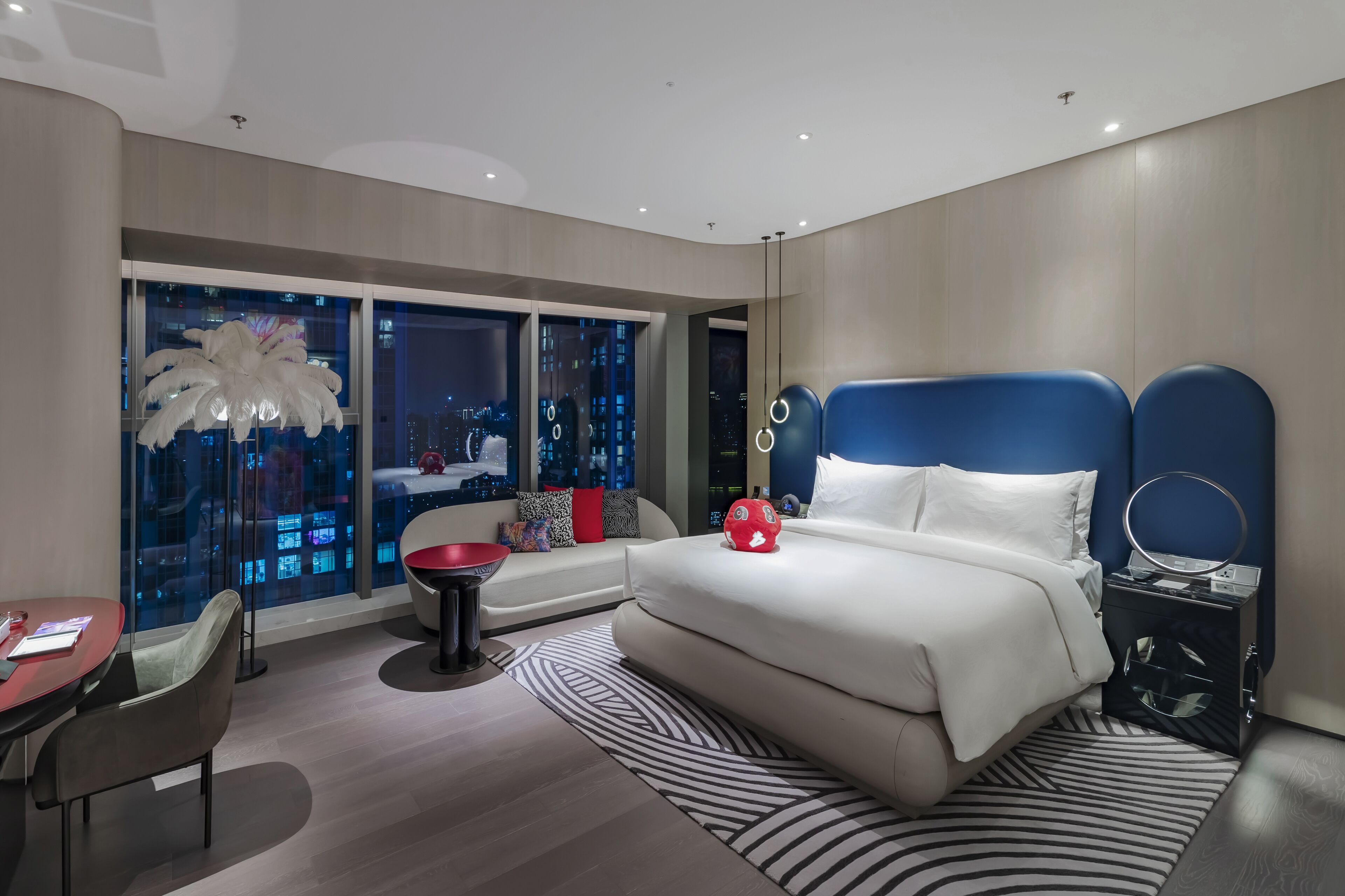 A modern hotel room with a large bed, a desk, a chair, and a view of the city at night.