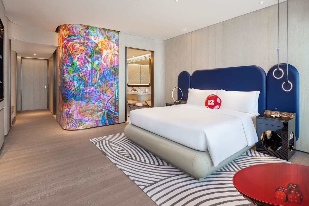 A modern hotel room with a large bed, a bedside table, a red circular table, and a colorful abstract wall.