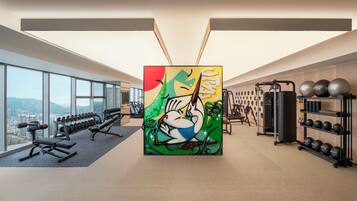 A modern gym with exercise equipment, a large window with a city view, and a colorful abstract painting.