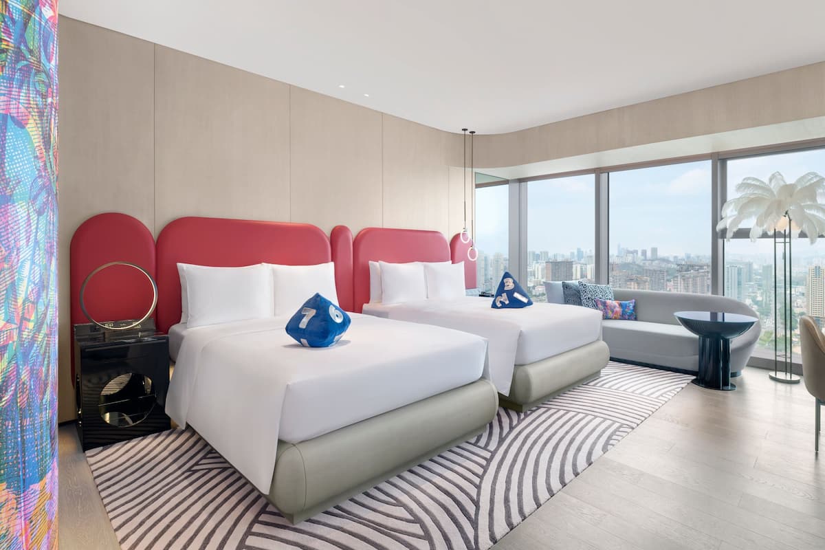 A modern hotel room with two beds, a sofa, a large window with a city view, and a patterned rug.