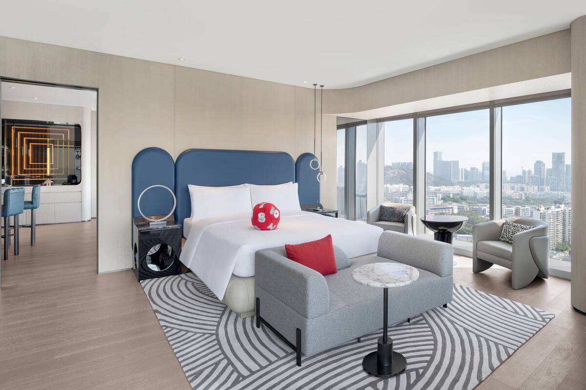A modern hotel room with a large bed, a grey armchair, a small round table, and a city view.