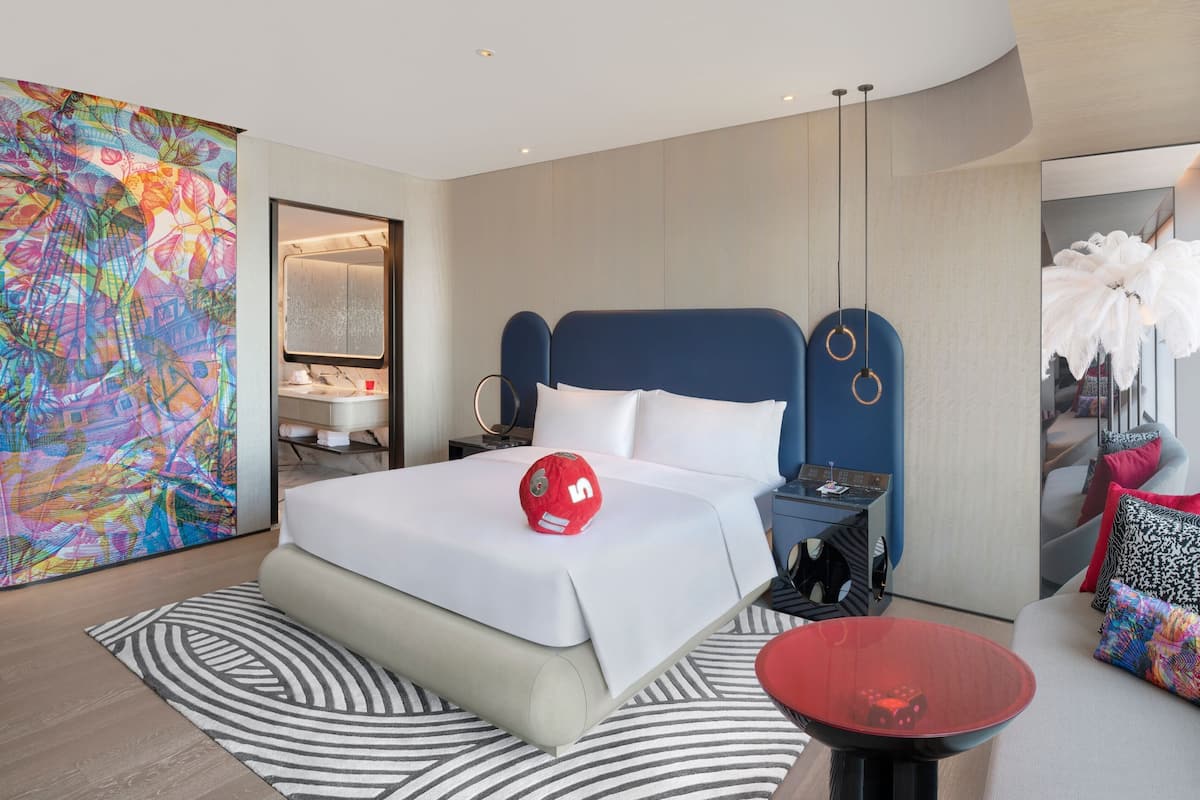 A modern hotel room with a large bed, a bedside table, a red decorative item, and a colorful abstract painting on the wall.