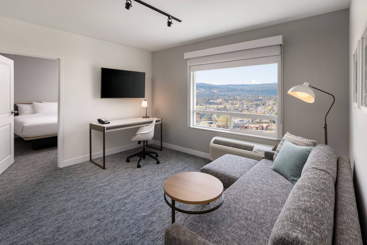 A modern hotel room with a large window, a gray sofa, a round table, a desk with a chair, and a bed.