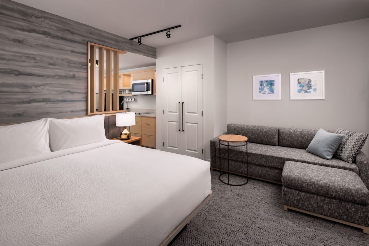 A hotel room with a large bed, a gray sofa, a small round table, a microwave in the background, and two framed pictures on the wall.