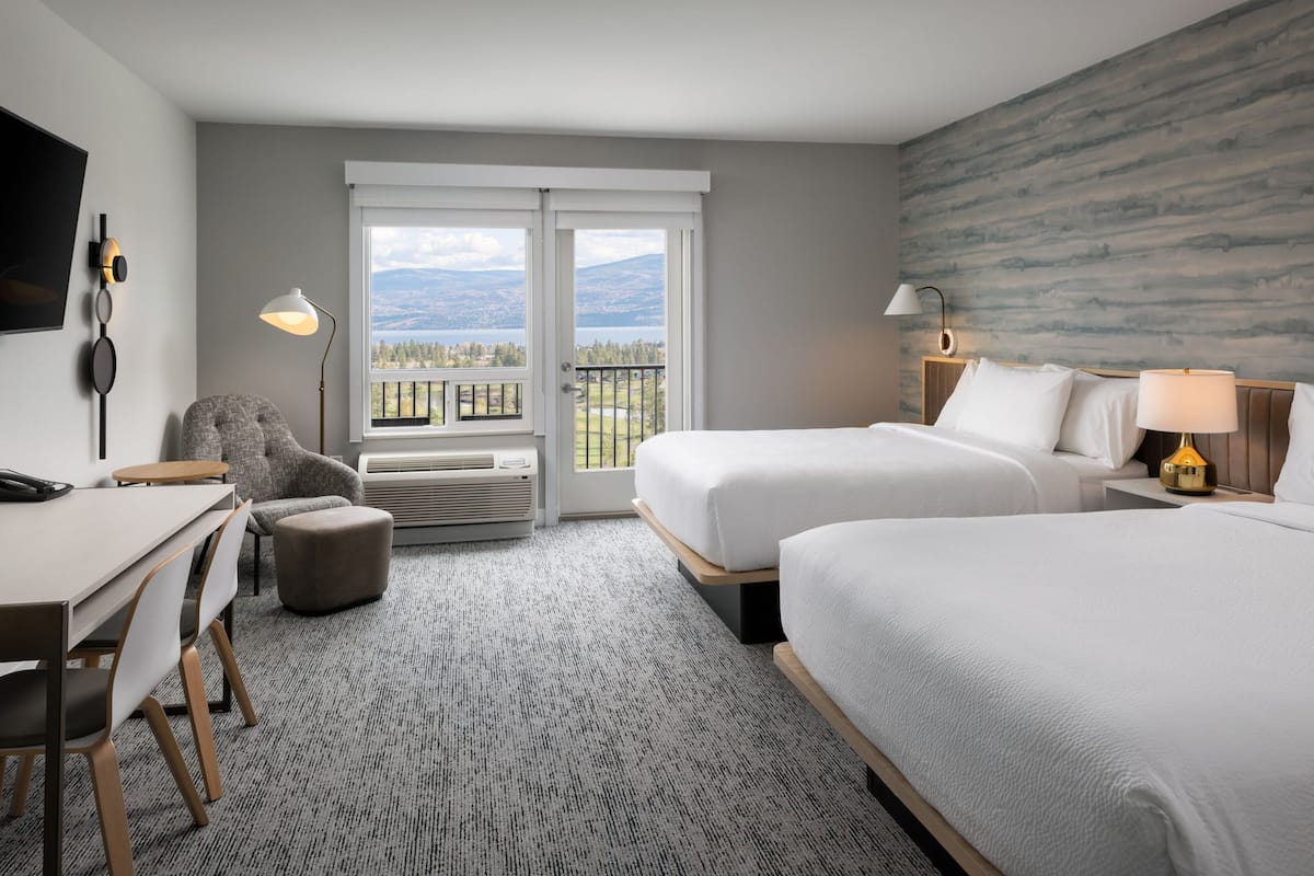 A hotel room with two beds, a desk, a chair, a balcony with a view of mountains, and a mounted TV.