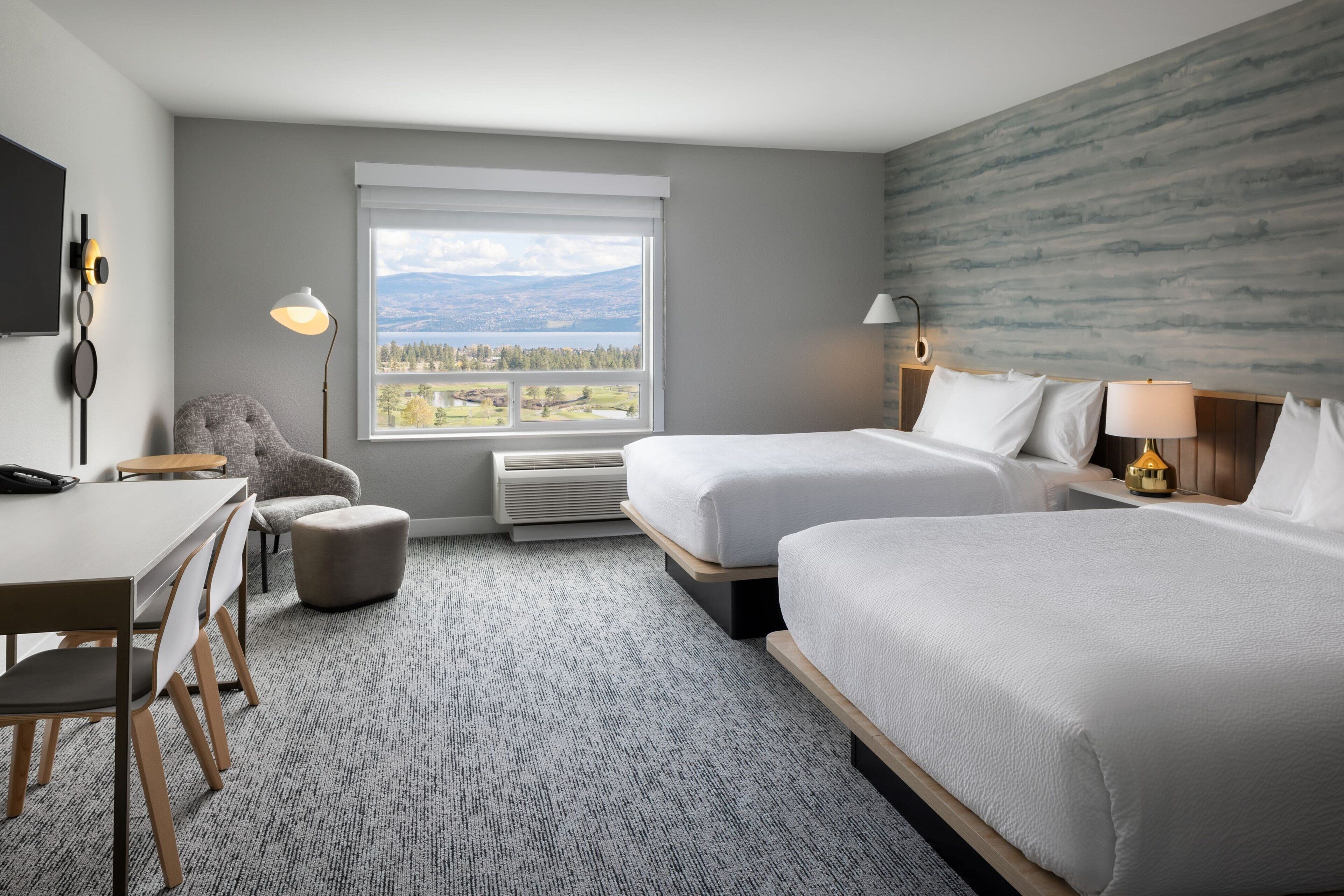 Une chambre d’hôtel avec deux lits, un bureau et une chaise. Une grande fenêtre offre une vue sur les montagnes et les arbres.