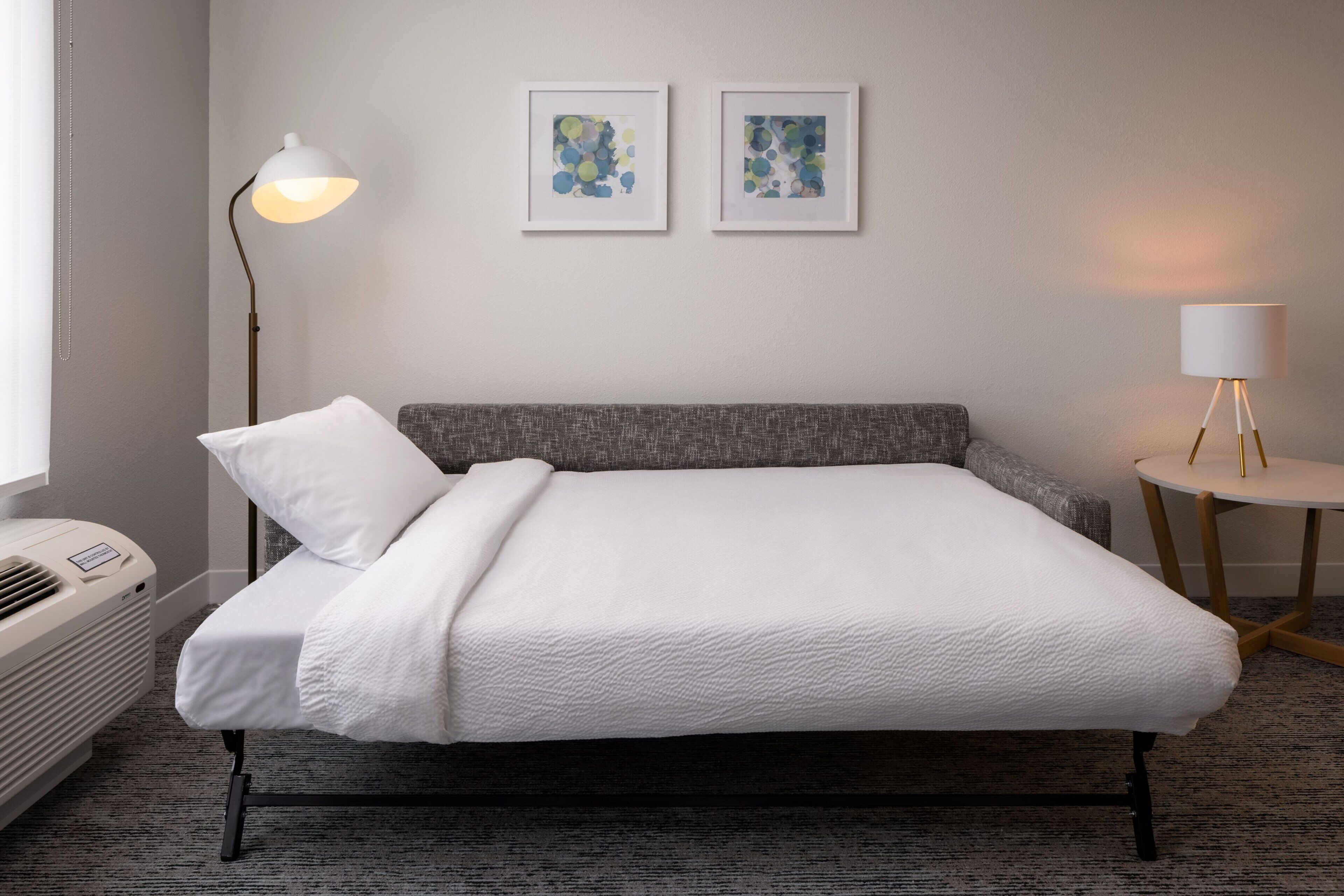 A neatly made bed with a white comforter and pillows, a gray upholstered headboard, a standing floor lamp, a small round table with a white lampshade, and two framed abstract art pieces on the wall.
