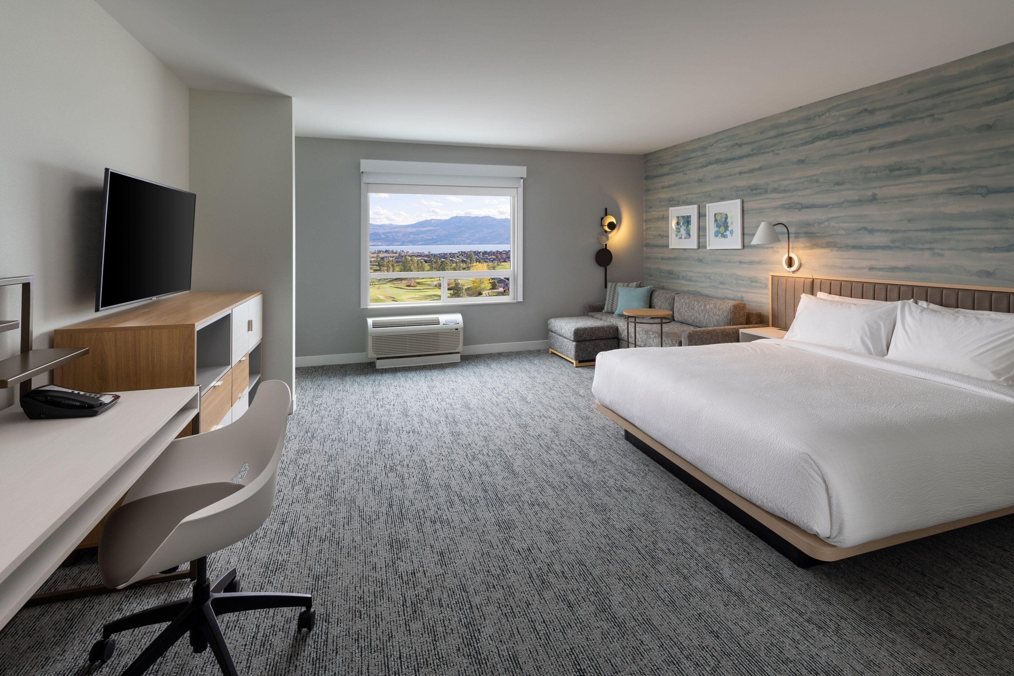 Quarto de hotel com cama, escrivaninha e cadeira, televisão, janela com vista para montanhas e luminária na parede.