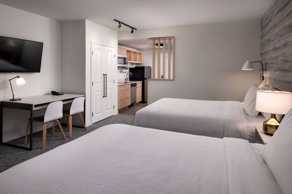 A hotel room with two beds, a desk, a chair, a TV, a microwave, and a small kitchenette.