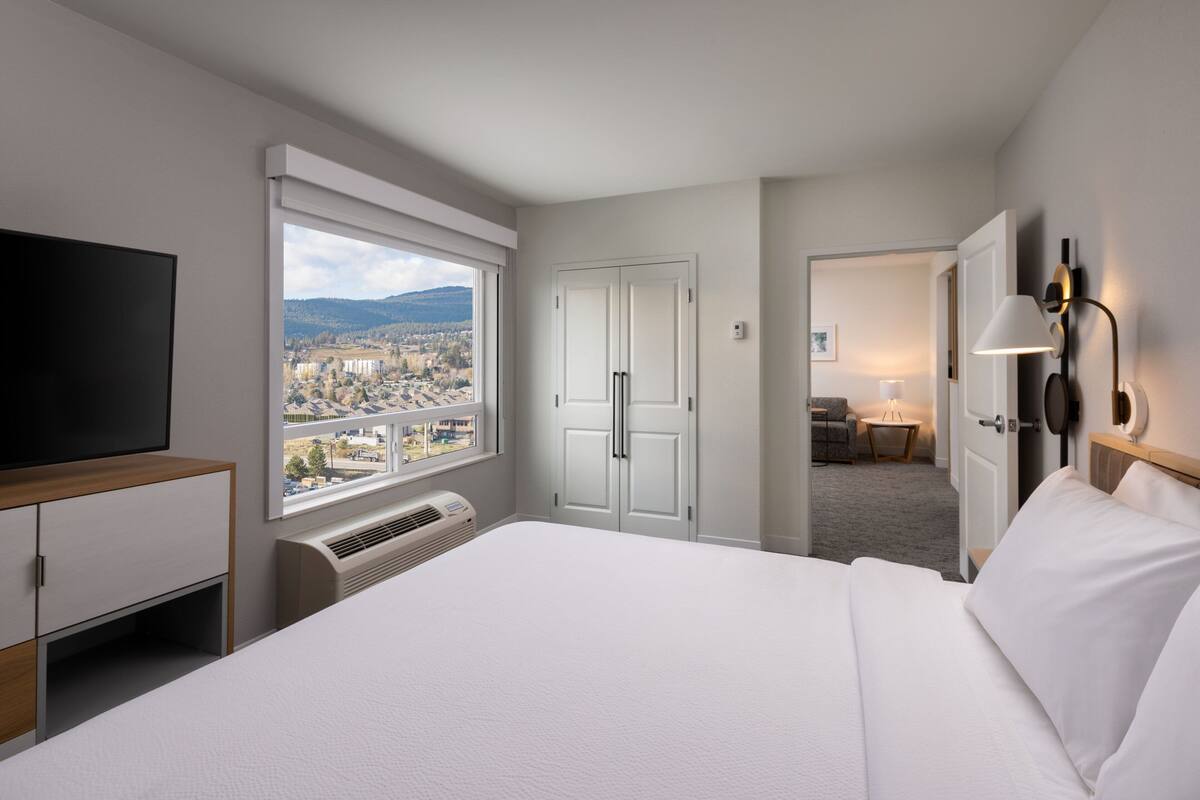 A hotel room with a large bed, a television, a wardrobe, and a view of the city through a large window.