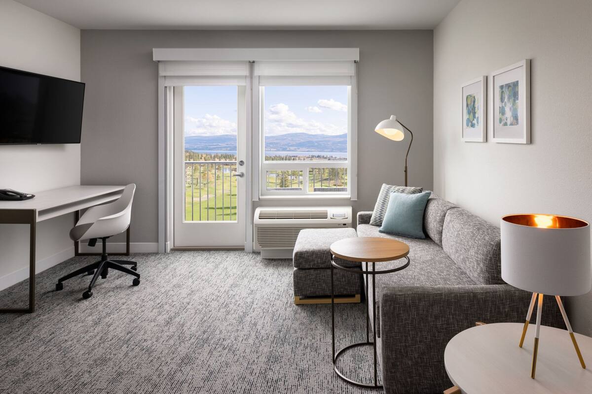 A modern hotel room with a desk, a grey sofa, a round table, a standing lamp, and a large window offering a view of the outdoors.