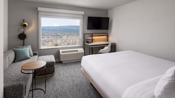 Habitación de hotel moderna con una cama grande, un sofá, un escritorio y vistas a la ciudad a través de un ventanal amplio.