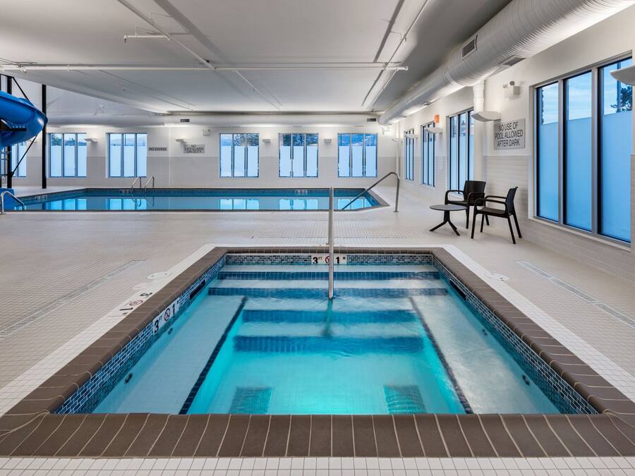 An indoor swimming pool with clear lane markings, a diving board, and a seating area with chairs.