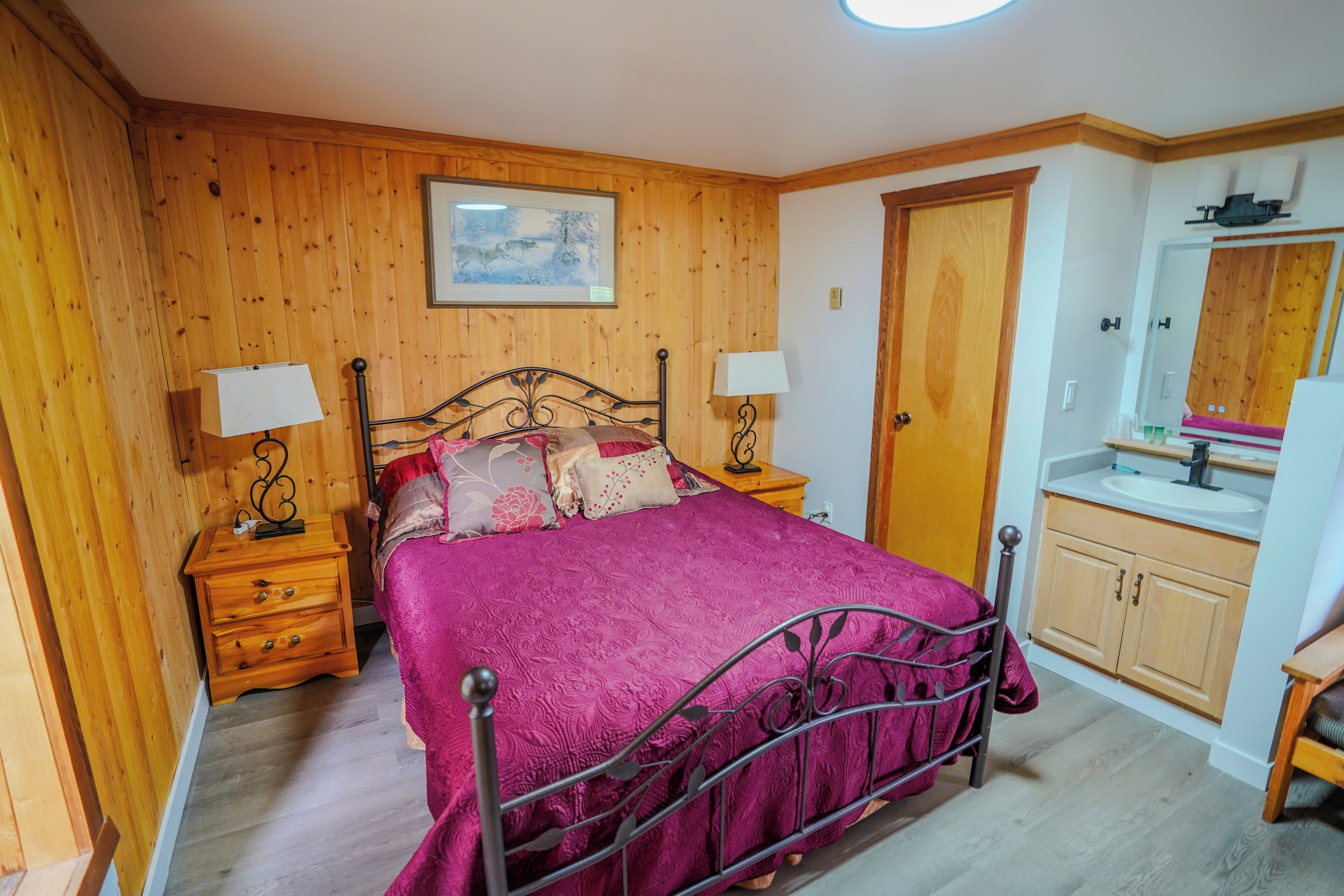 A bedroom with wooden walls, a metal bed frame, a wooden nightstand, a bedside table, a wooden door, a bathroom with a sink and mirror.