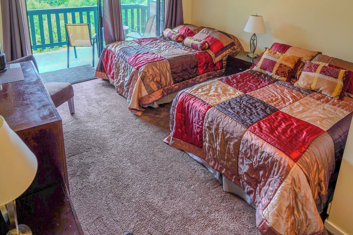 A bedroom with two beds, a desk, and a chair. There is a balcony with a view of greenery.