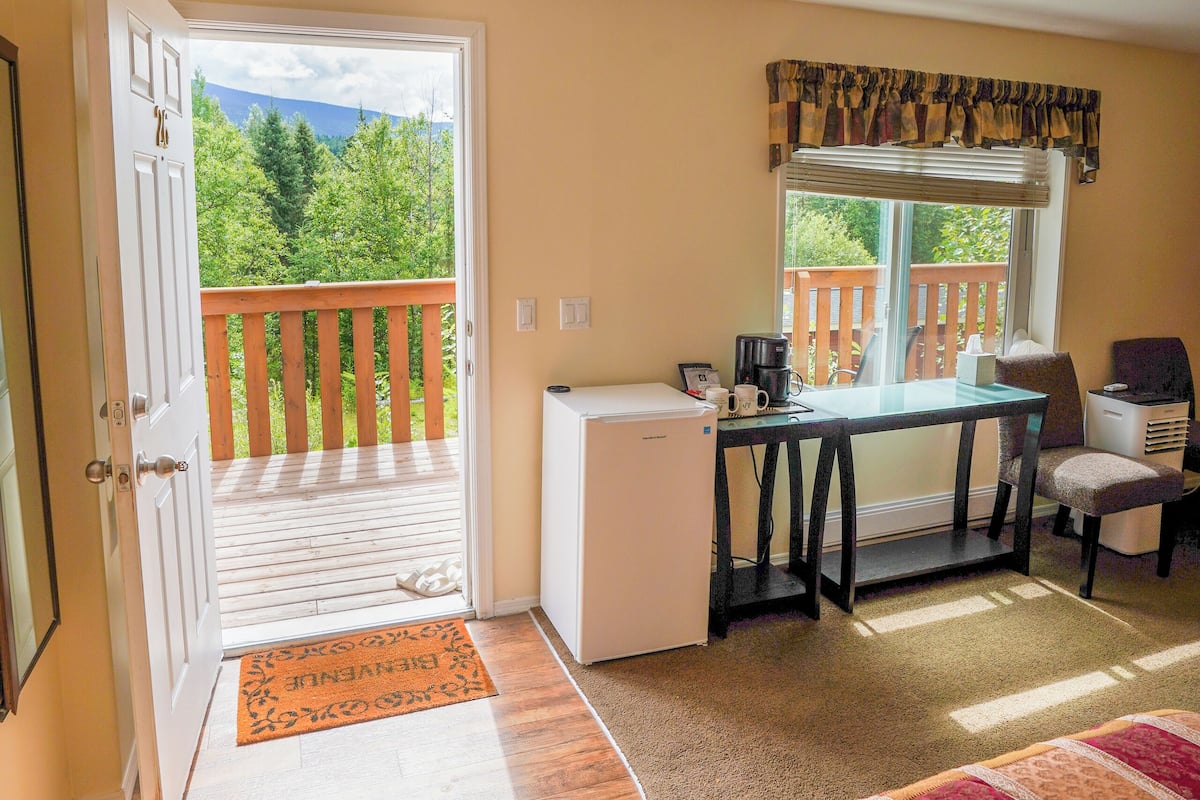 A room with a balcony, a small desk with a chair, a bed with a patterned bedspread, and a window with a view of greenery.