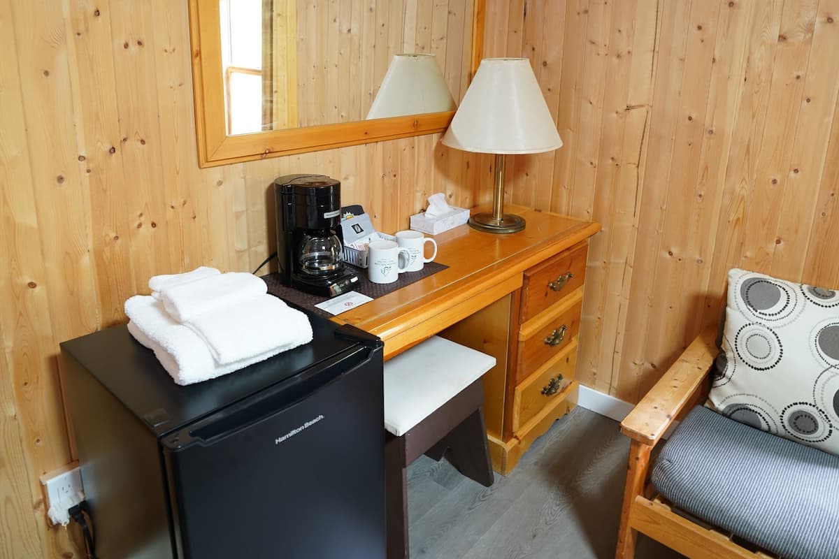A wooden-paneled room with a desk, a black refrigerator, a coffee maker, a lamp, a chair with a patterned cushion, and a trash bin.