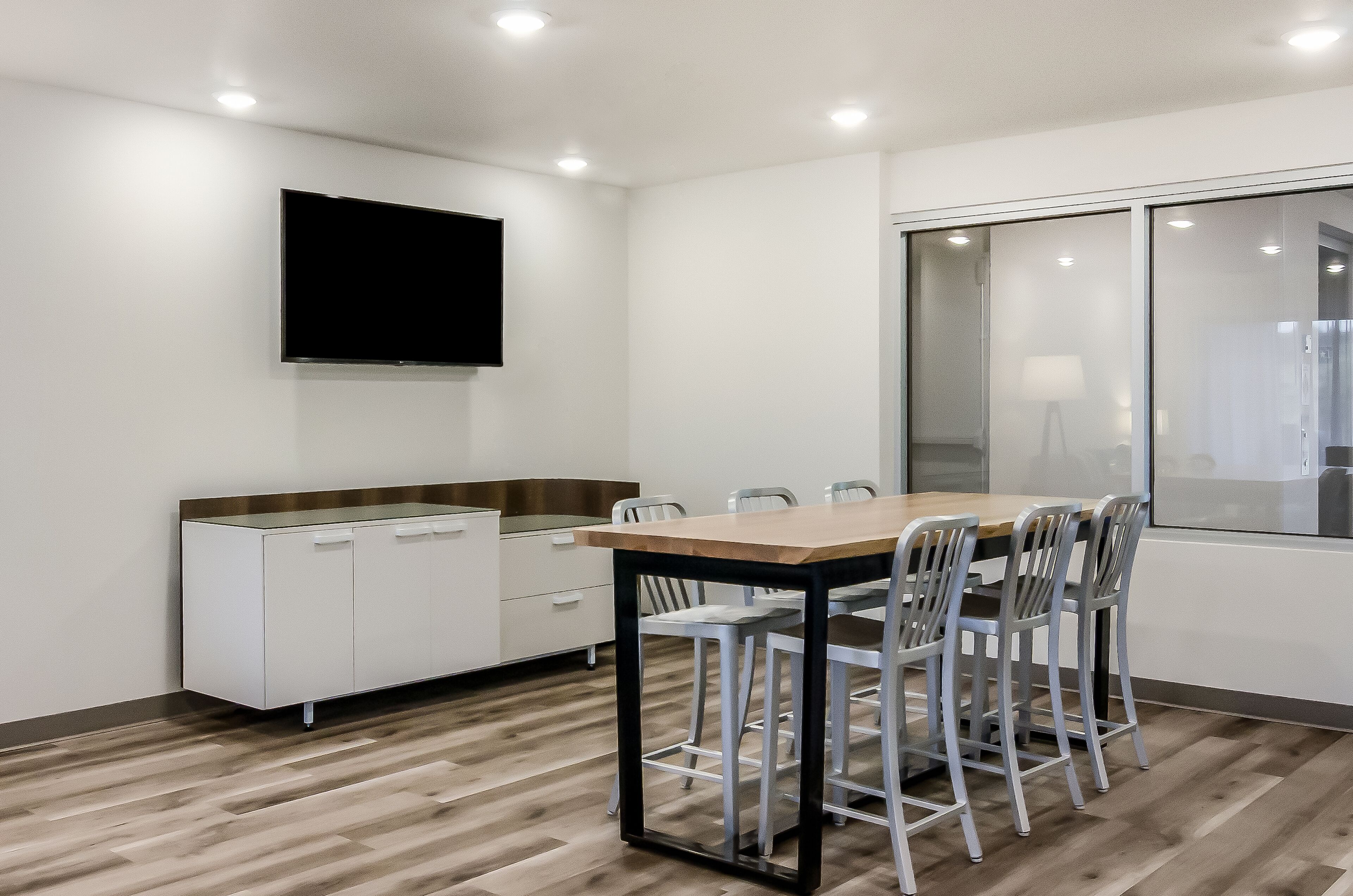 A modern kitchen with a wooden table and chairs, a white cabinet, and a flat-screen TV mounted on the wall.