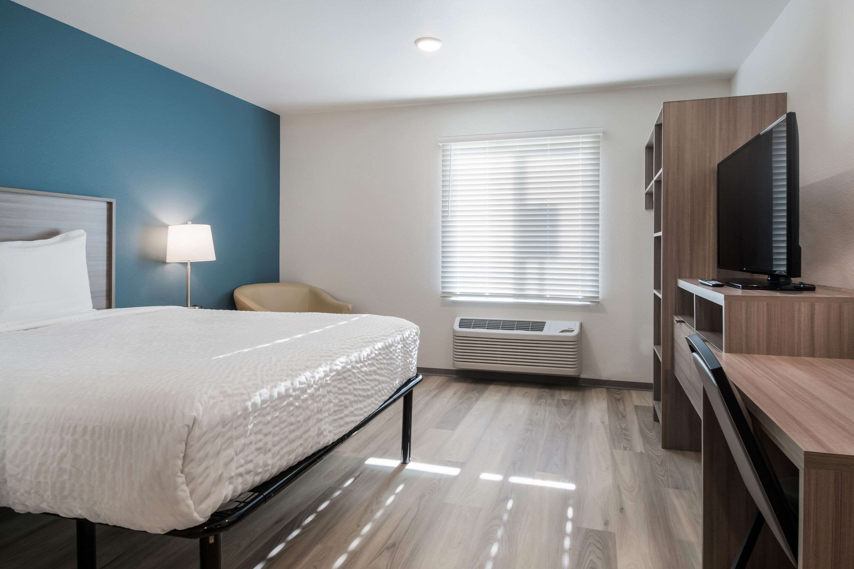 Una habitación de hotel con una cama, televisión, sillón y ventana con persianas.