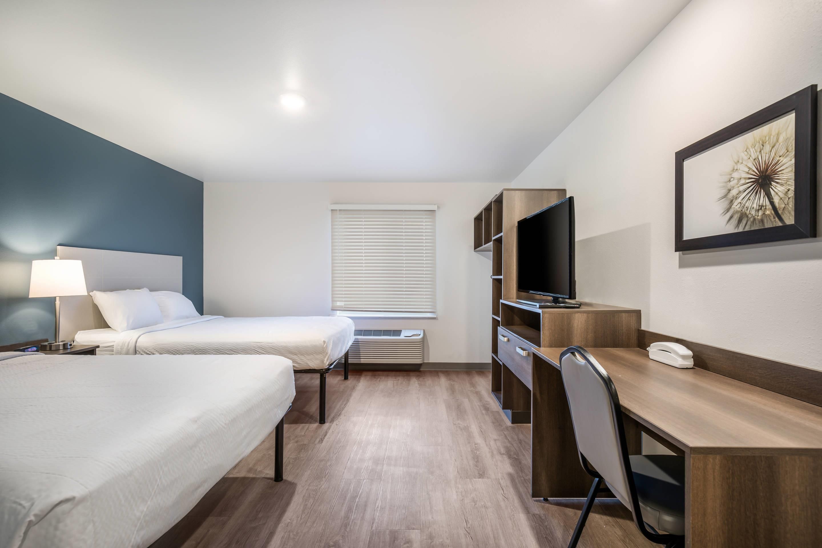 Habitación de hotel con dos camas, un escritorio con silla, un televisor y una ventana con persianas.