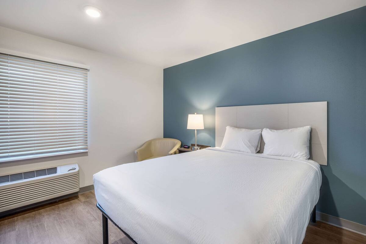 A hotel room with a large bed, a bedside table with a lamp, a chair, and a window with blinds.