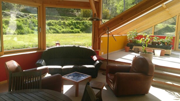 Een gezellige kamer met een bank, stoelen, een tafel en grote ramen met uitzicht op een tuin.