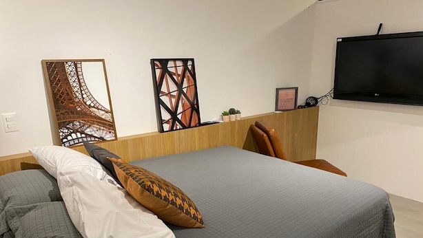 Una cama bien hecha con una colcha gris y almohadas, un cabecero de madera, un televisor montado en la pared y decoración mural.