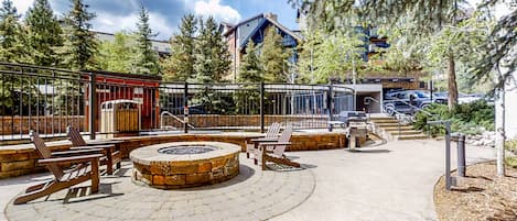 Relax in a cozy seating area surrounded by trees and a modern building, featuring a fire pit and Adirondack chairs.