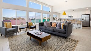 A spacious living room with a sofa, armchairs, and a coffee table. A kitchen with modern appliances and a yellow pendant light is visible in the background.