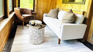 A cozy room with a wooden interior, a white sofa with cushions, a leather armchair, and a patterned rug. - Spacious Fly Creek Retreat – Game Rm, Creek Views & Minutes to Cooperstown & DP (Fly Creek)