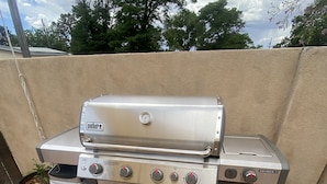 A Weber Genesis II portable grill with four burners, a thermometer, and a side burner, placed on a concrete patio with potted plants nearby. - Garden Patio Studio near Bosque, Old Town (Albuquerque)